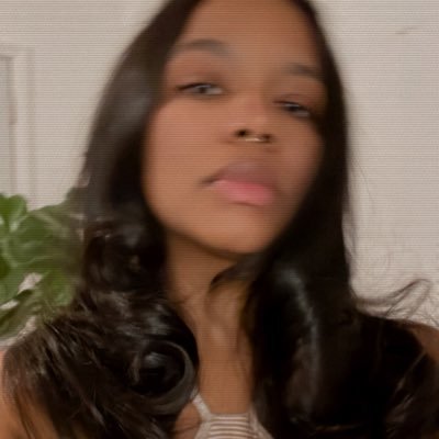 Elizabeth, a brown skinned person, wears her hair straight and appears blurred and distorted. In the background there is a plant.