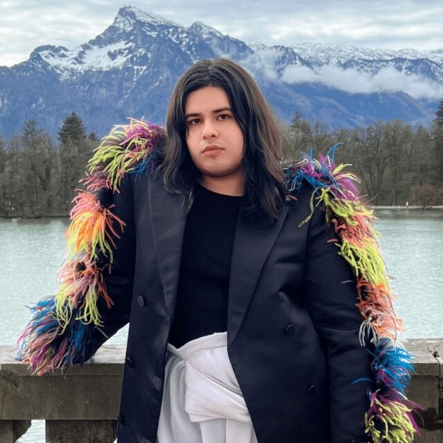 Pictured is artist Kira Xonorika. They are a person with dark brown, shoulder-length straight hair with a slight wave, parted in the middle. They are standing outdoors, leaning on a stone balustrade overlooking a serene lake and a mountainous landscape, with bare trees surrounding them. They wear a black top and a light black coat adorned with vibrant, multi-colored feathers cascading down the sleeve, and white high-rise, flowy, linen-like fabric that ties around the waist. In the distance behind them, snow-capped peaks and a cloudy sky form a picturesque backdrop.