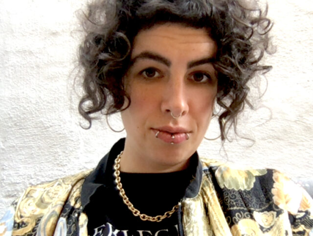 On the image is a femme presenting German-Iranian person with light skin and ear-length curly dark hair and dark eyes, with several piercings and wearing a golden, patterned jacket over a black shirt with a type print, as well as a golden chain necklace.