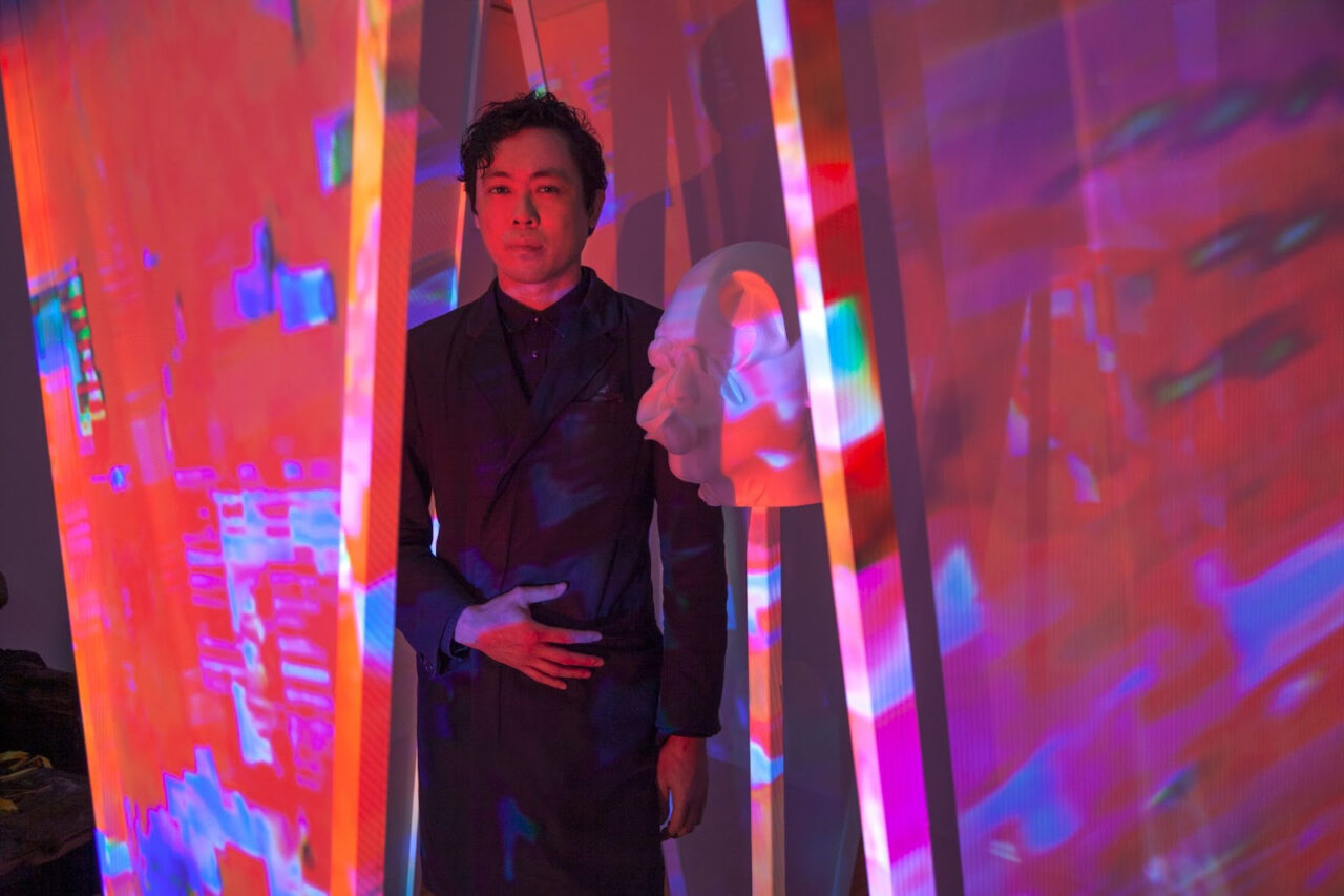 Photograph of Chinese-American man standing inside an artist-built structure next to a 3D printed sculpture on a wooden stick. The light of a red video projection with blue digital artifacts washing over the entire structure, figure, and sculpture. He has short curly hair, wearing a black long jacket and black button up shirt.