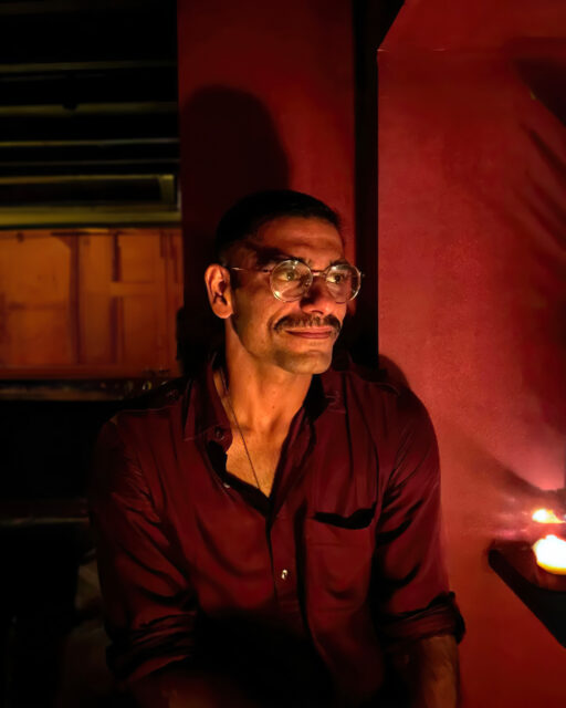 Pictured is artist Sahej, an indian man, in a dimly lit, red-toned room. Sahej wears a dark red/brown shirt that is opened at the collar and thin framed glasses. He has a neatly shaven moustache and light beard, and dark brown hair.