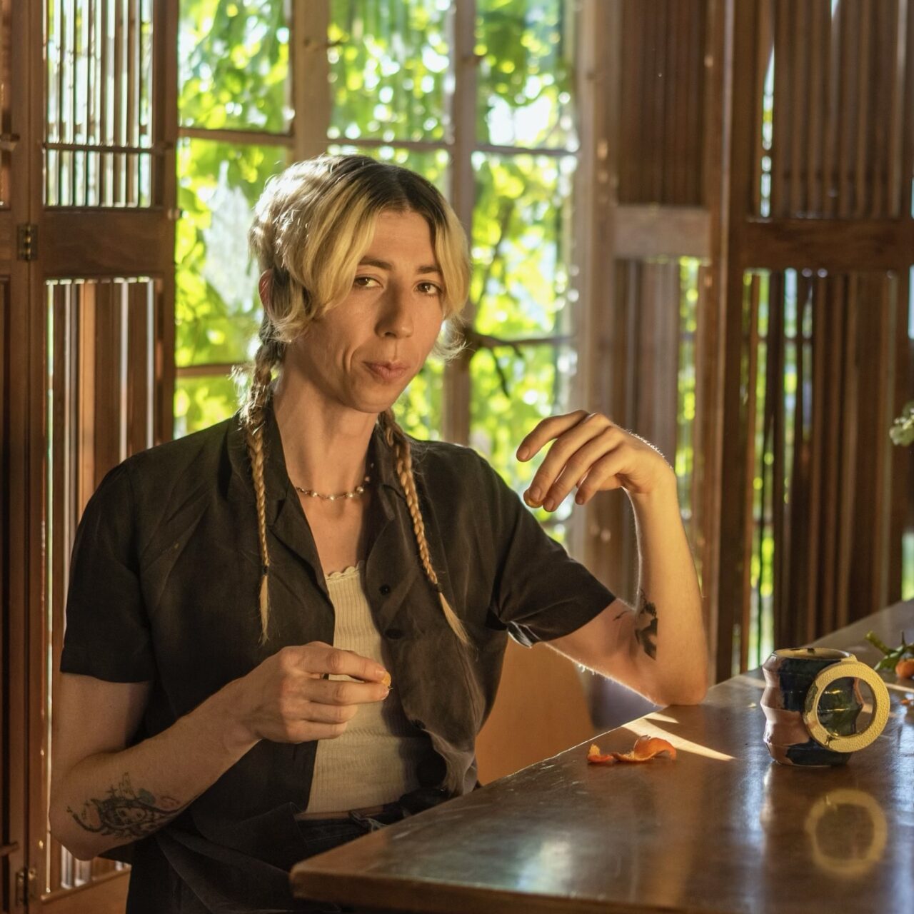 A white tattooed trans woman sits at a kitchen table backlit by a green passionflower vine in the window. She has blond center-parted short hair, with long twin braids draped over the front of her open charcoal button up shirt. She looks into the camera while eating a tangerine.
