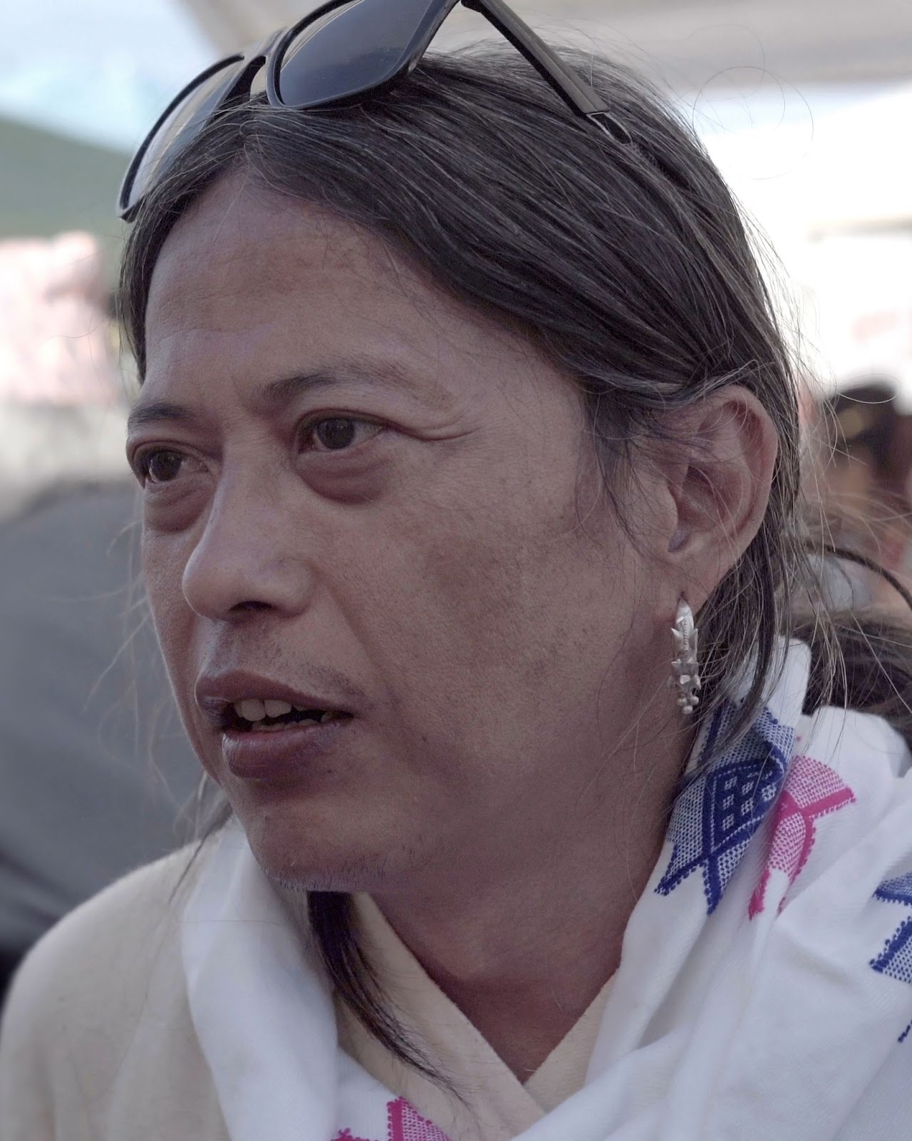 An indigenous Yakthung person with long hair, silver earrings, traditional white scarf with blue and pink star patterns.