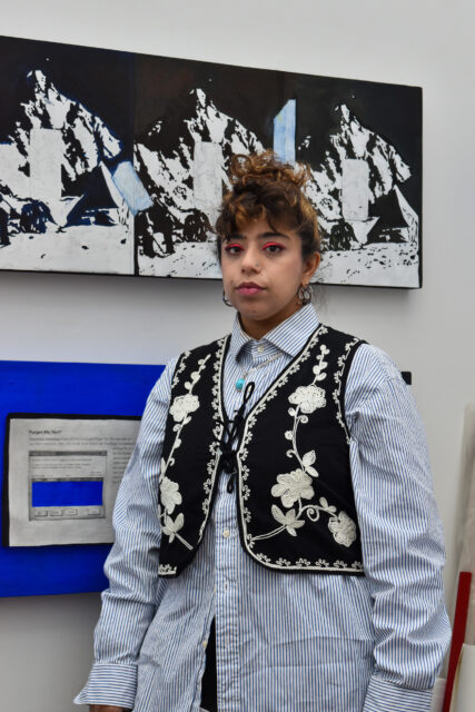 Pictured is artist Umber Majeed. She stands against a white wall displaying two pieces of artwork. They have brown hair styled in an updo with curly bangs, and are wearing bright pink eyeliner, a nose ring, and thick hoop earrings. She is dressed in a light blue-and-white striped button-down shirt layered under a black vest featuring white floral embroidery and a lace-up front. The artwork behind her includes a black-and-white mountain scene and a blue canvas with a vintage computer window graphic.