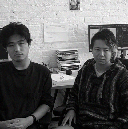 Black and white photo of artists, Yang Wang (left) and Zhenzhen Qi (right). They are both seated in their studio in front of a white brick wall.