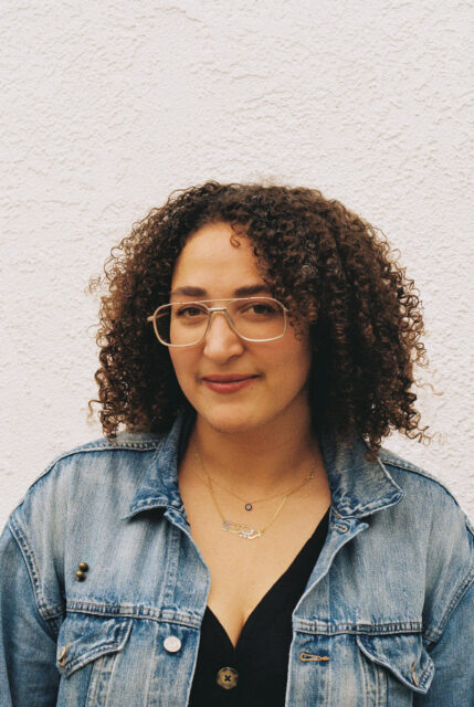 A person with light brown skin and medium brown curly hair is in front of a white stucco wall. Smiling at the camera they are wearing a black top, blue jean jacket, gold-rimmed glasses, and an evil eye and Arabic nameplate necklace.