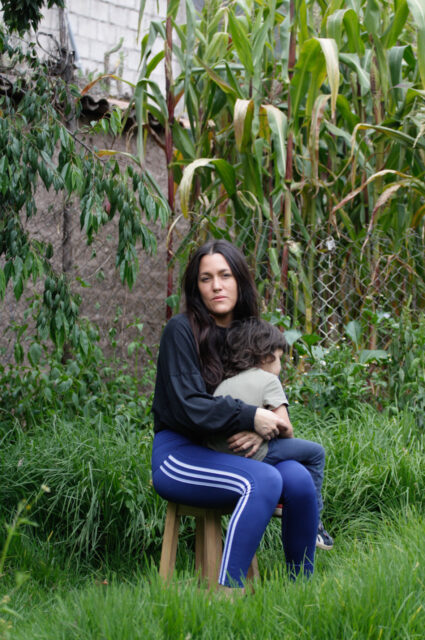 Pictured is a light skin woman with long dark brown hair seated on a light wooden stool carrying her child, who is facing away from the camera. The woman wears a long-sleeve black blouse and blue sweatpants with three white stripes that go up and down the leg. They are seated in a lush green backyard.