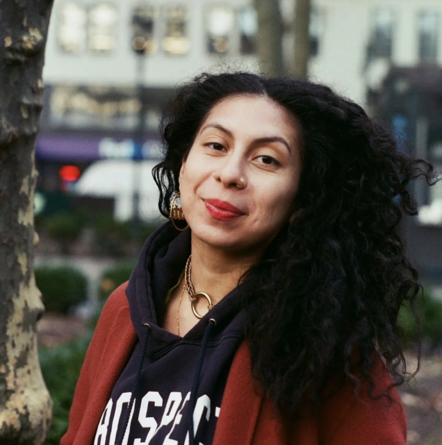 Pictured is artist, dre jácome, a brown femme with big black curly hair with gold hoops and nose rings smiles wearing a terracotta coat over a blue hoodie. dre smiling outdoors in NYC streets.