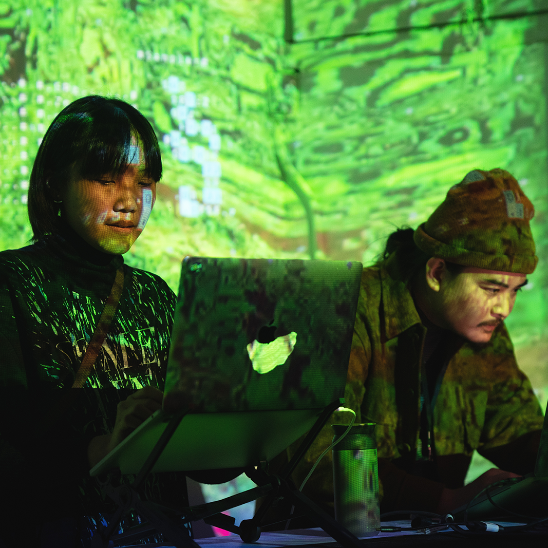 A profile photo of Nitcha(left), a Southeast Asian woman wearing a black t-shirt with a white graphic. She has short, straight hair with bangs. Kengchakaj (Keng) (right) is a Southeast Asian man wearing a beige beanie and jacket. They’re standing and typing on their laptops with green graphic projection lights on their faces and backgrounds.