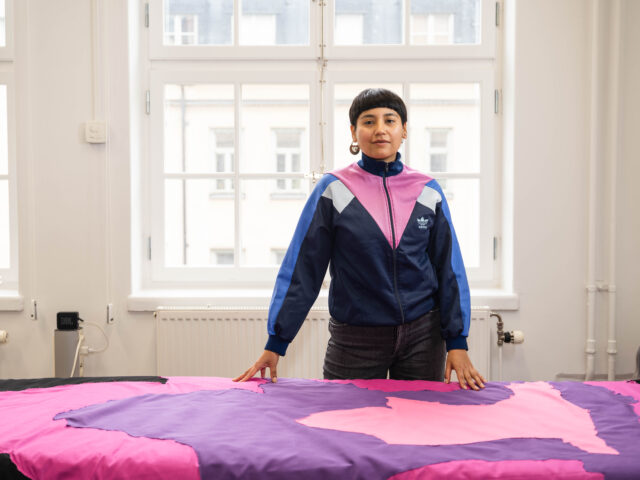 Standing person with brown skin, short black hair, traditional Mapuche earring, blue, light blue and pink sweatshirt with zipper in the middle. In front of their is a part of a textile piece in light pink, dark pink, purple and black. The piece is on a table where the artist rests their hands.
