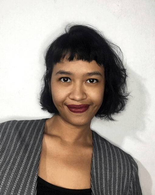 Pictured is artist Syaura, a brown-skinned Indonesia Woman, with dark brown hair wearing blunt bangs and wavy bobbed hair that is mid-neck length. She wears burgundy color lipstick and a pin-striped jacket over a black top.