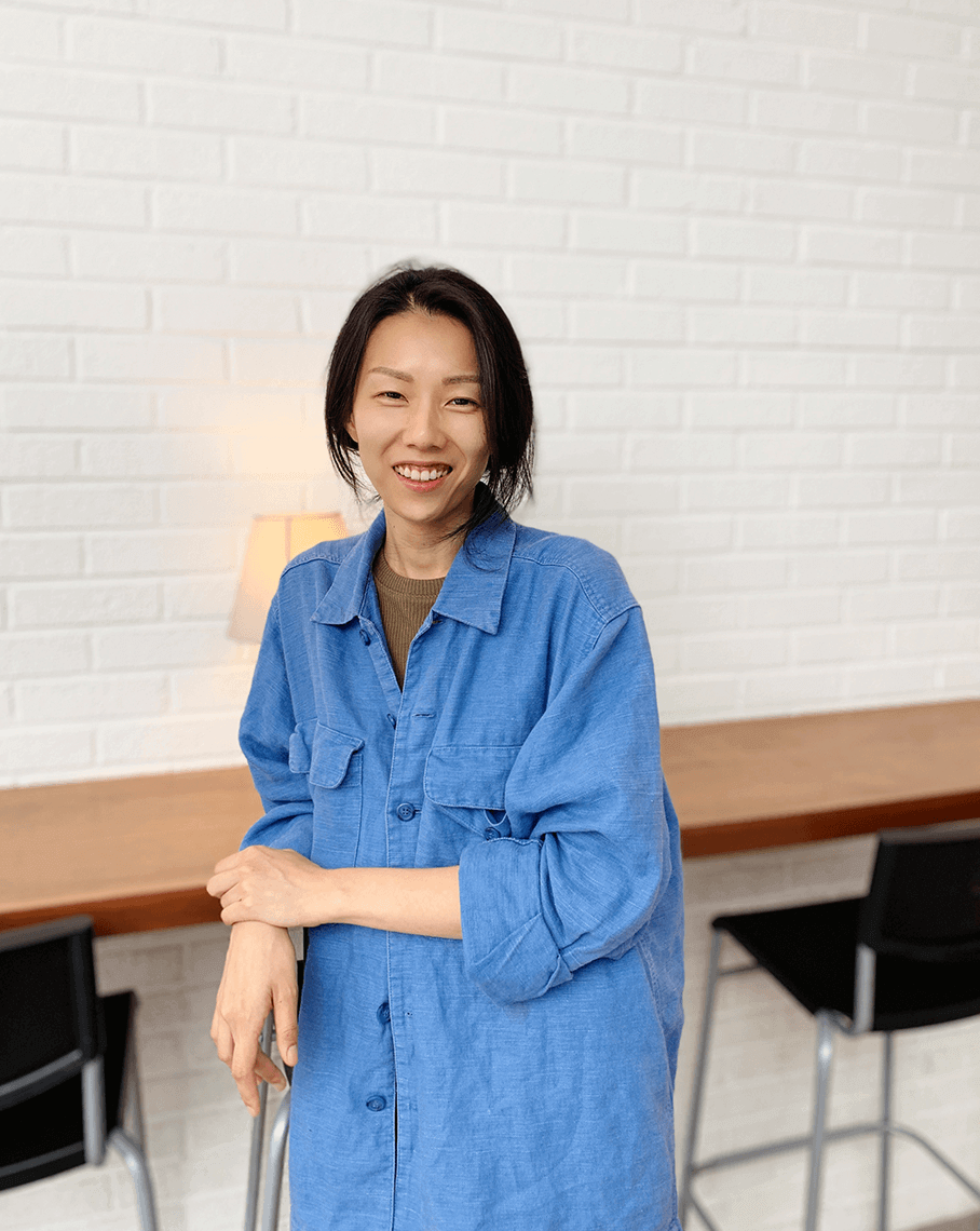 Pictured in artist Jungwong Seo, a Korean woman with dark auburn hair who wears a oversized blue linen buttoned shirt. She stands leaning in front of a chair against a white brick wall.