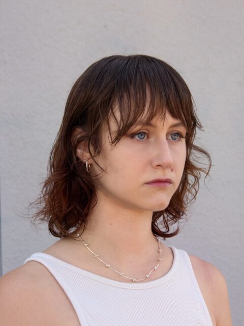 Pictured is artist Aurora Mititelu, a Romanian woman. She has light skin, blue eyes, and shoulder-length brown hair with bangs. They are looking off-camera to the right with a neutral expression. They have a beauty mark on their cheek and are wearing a white ribbed tank top, small silver hoop earrings, and a thin silver necklace with small beads. The background is a plain, light grey wall.