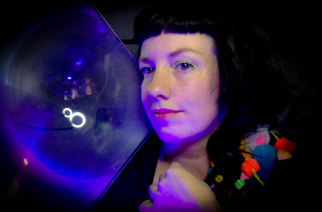 Portrait of a dark haired caucasian woman in a dark room. Her hair is black and cut with a short black fringe, her eyes are accentuated with yellow eyeliner and her lips are purple red. She holds a projector lens that shows a vague outline of the reflection of her face projected into infinity.