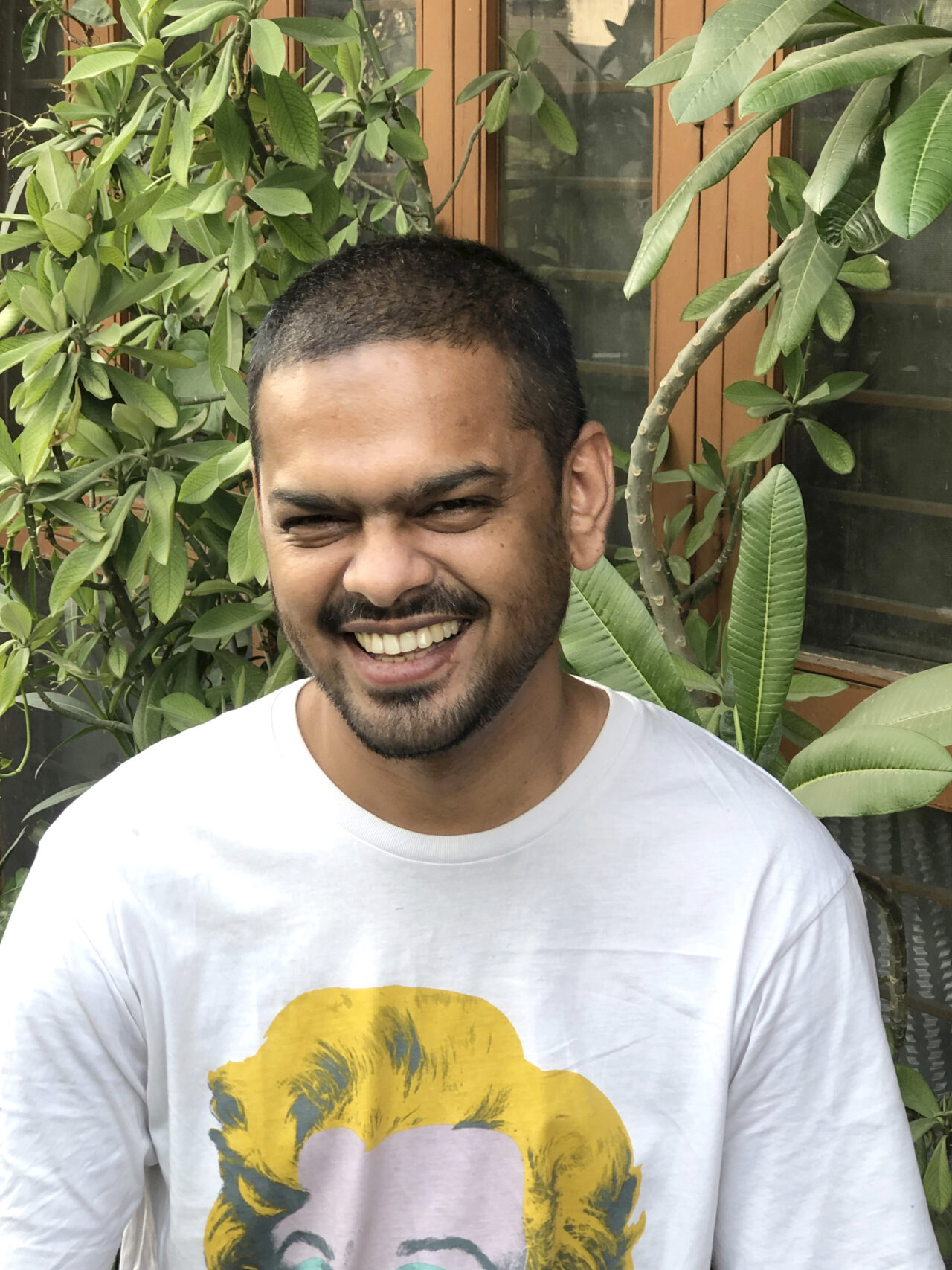 Pictured is Parabartana Mohanty, a lighter skin Indian man, with buzzed hair, and a trimmed mustache and beard. He wears a white t-shirt with a print of one of Andy Warhol's Marilyn Monroe Diptychs. He is smiling with teeth baring, and standing in front a wall of green leaves.