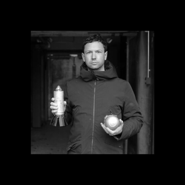 The picture shows a self-described young-man with short hair and a five o'clock shadow holding a cylindrical object and a sphere, looking right into the camera with a straight face. The picture is in greyscale.