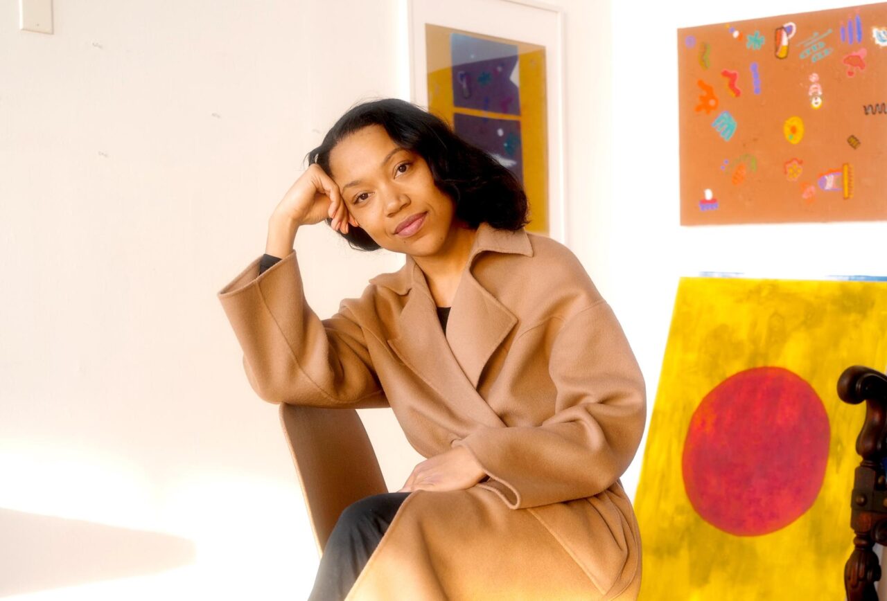 A light brown woman with medium-length wavy and dark hair, Ari Melenciano, sits in her studio in front of a few colorful and abstracted art hanging on the wall and a large painting leaning against the wall . Image courtesy of the artist