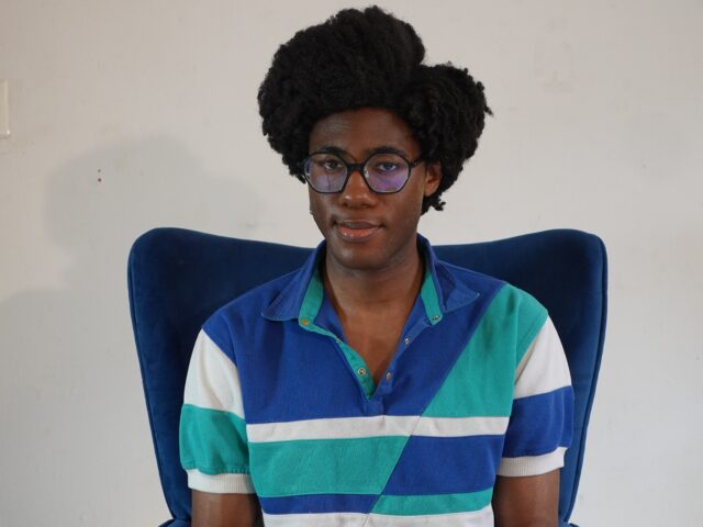 Avery Alex Beige seated in a high-backed navy blue velvet chair against a plain white wall. They have voluminous natural hair and wear black, large-framed, geometric glasses. They are wearing a short-sleeved, color-blocked polo shirt featuring blue, teal, and white sections. They are looking directly at the camera with a subtle, gentle expression.