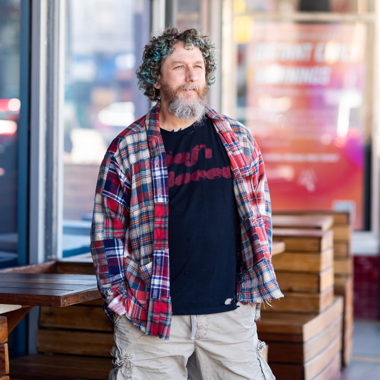 A middle-aged man squints from the sun. He has pink skin, light-brown, mid-length curly hair with blue tips, and a long curly beard with a greying goatee. He’s wearing a black t-shirt printed with red text, a patchwork red flannel shirt, and khaki pants. Behind him is outdoor seating.