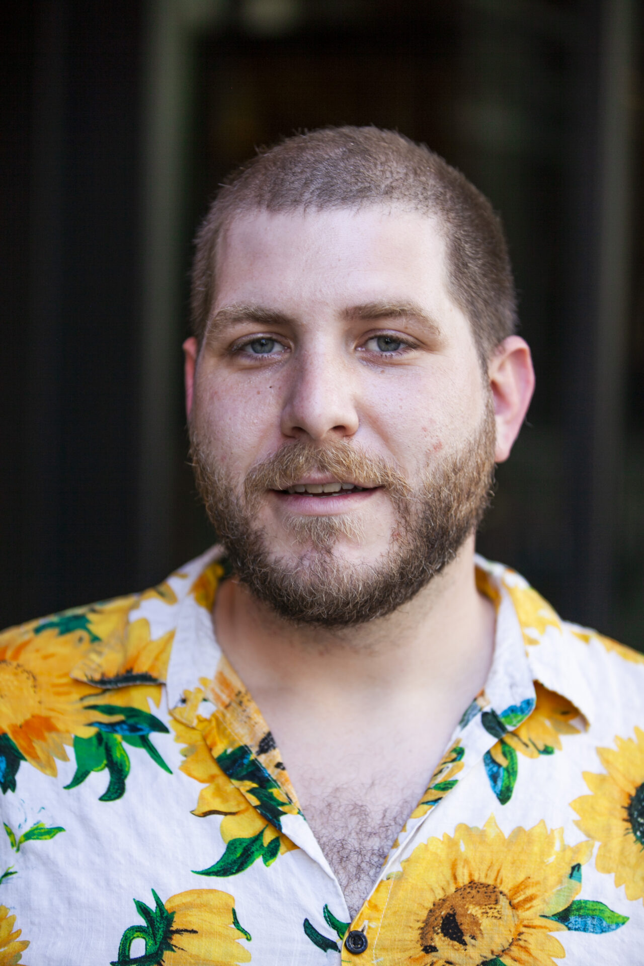 Pictured is artist Roo, a fair skin white-passing indigenous person with buzzed hair, trimmed moustache, beard. Roo wears a button up shirt with patterns of bright yellow flowers.
