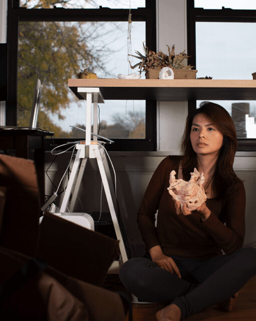 Pictured is Doreen Chan, who sits on a wooden floor, and holds a small craggy rock-shaped sculpture in her left hand. She has warm auburn, brown hair that swoops down below her shoulders. She is seated by a work table, in front of two windows looking out to a park.