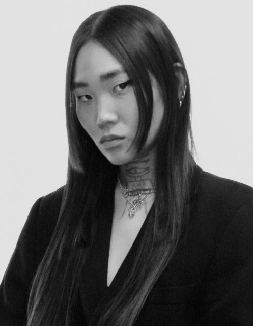Pictured in Black and White, is a korean-american person with dark straight hair parted in the middle, wearing a black deep-V jacket. Has a tattoo on neck.