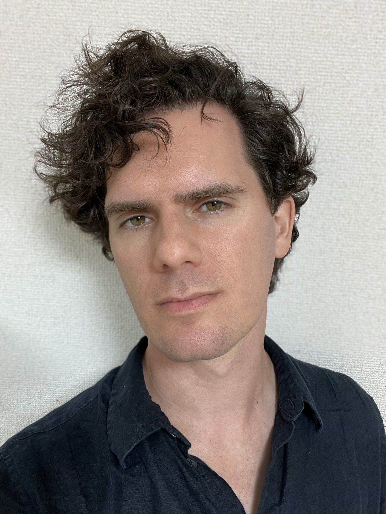 Self-portrait of artist Kyle McDonald. Mid-30s white male with short wavy brown hair, neutral face expression, clean shaven, collared black t-shirt, against a white background.