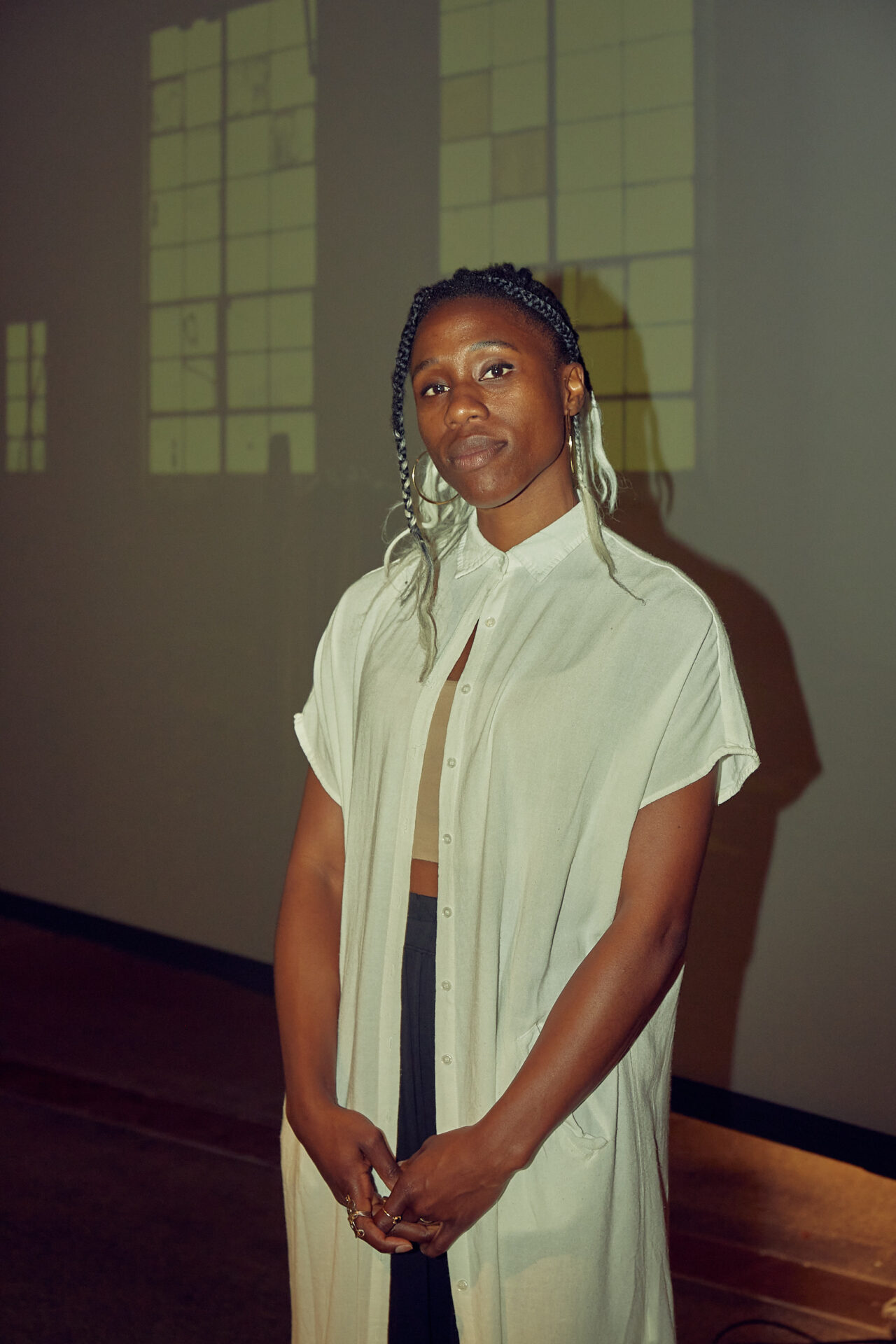Artist Mimi Ọnụọha standing in front of her video installation These Networks In Our Skin. She is a dark-skin nigerian woman who wears a white sheer top and braids.