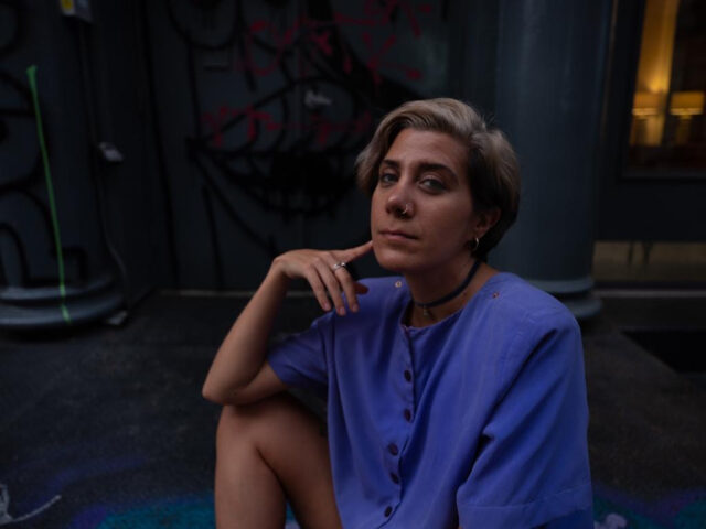 A profile image of a brown woman with short, light-brown side-swept hair wearing a denim blue buttoned top, sitting in front of a wall covered in black and red spray-painted street art between two dark grey columns.
