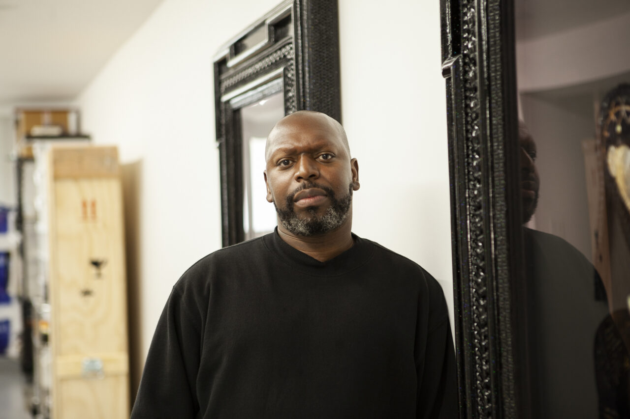 Portrait of Rashaad Newsome, a dark-skinned Black man with salt and pepper beard and moustache. He stares directly at the camera and wears a black sweater. Newsome stands in front of two dark wooden-framed mirrors on a white wall.