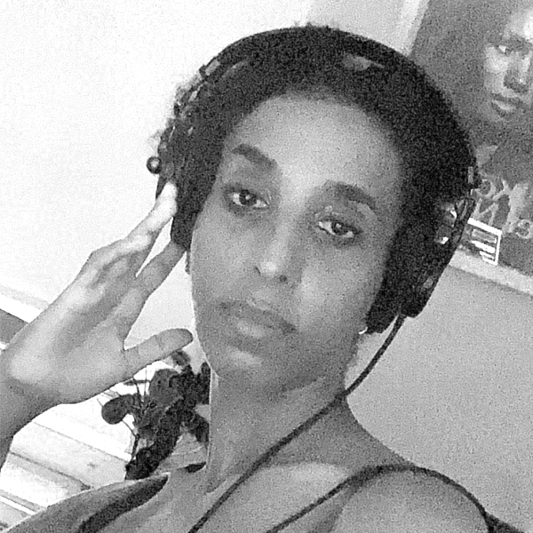 This is a black and white grainy photo of a close up of Rodan Tekle's face, she is a black woman with headphones on, one hand touching the headphone as she's looking straight into the camera with a serious but gentle gaze.