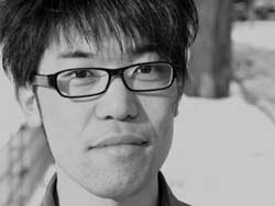 Pictured is Satoshi Morita, a japanese sound artist. He has short black hair with long bangs that cover his forehead and thick rimmed black glasses.