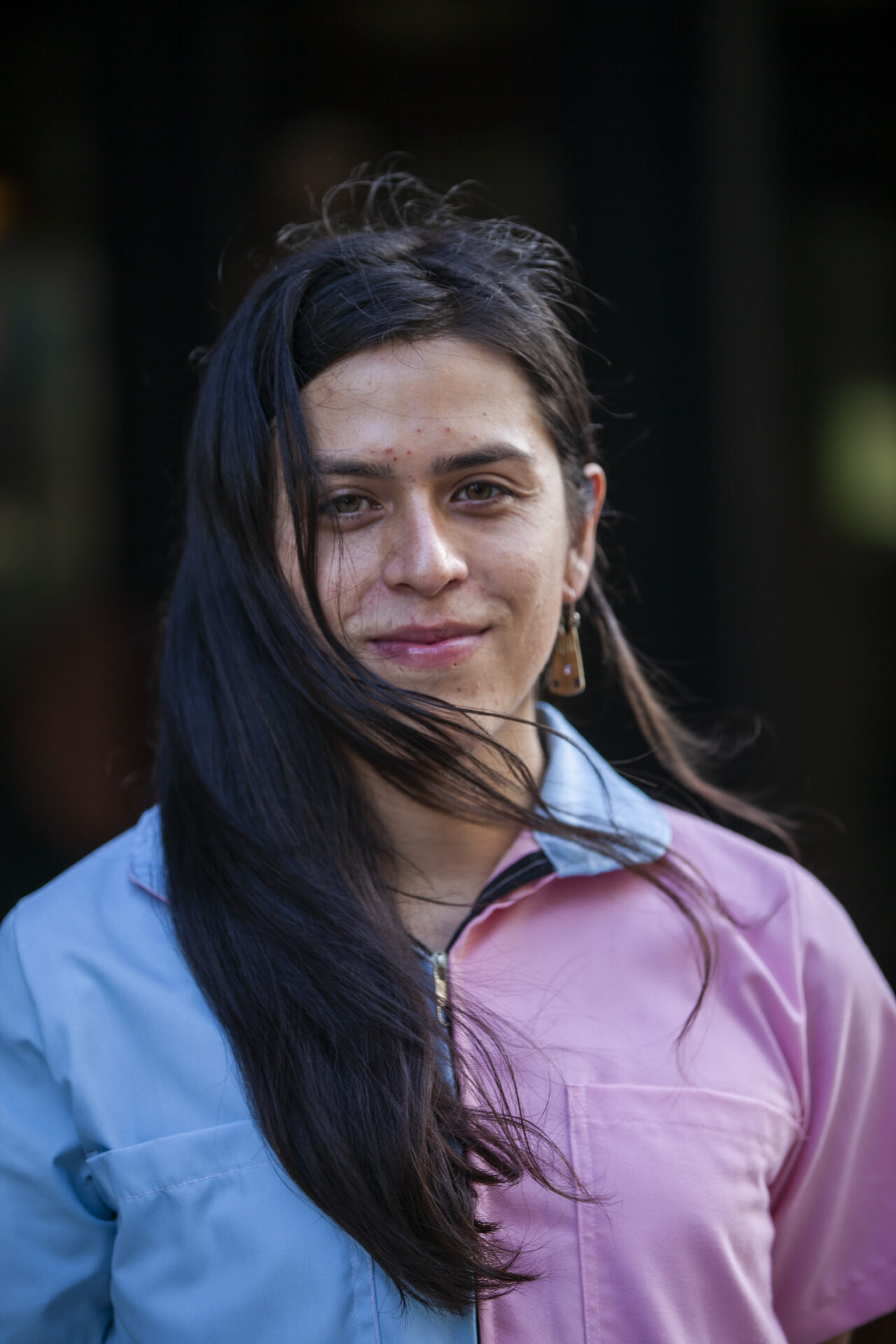 Pictured is an indigenous person with long dark brown straight hair and medium brown skin. they wear a jumpsuit that is light blue on one side and pastel pink on the other.