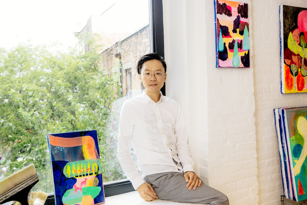 Pictured is artist, Taeyoon Choi. He is a light-skinned Asian man, with shiny dark black straight hair, slicked to the side. He wears clear round frame glasses, a white button up shirt, and grey linen pants. He sits in front of a window that opens to a lush green tree, and to his right and left are canvas paintings with bright colors against a white brick wall.