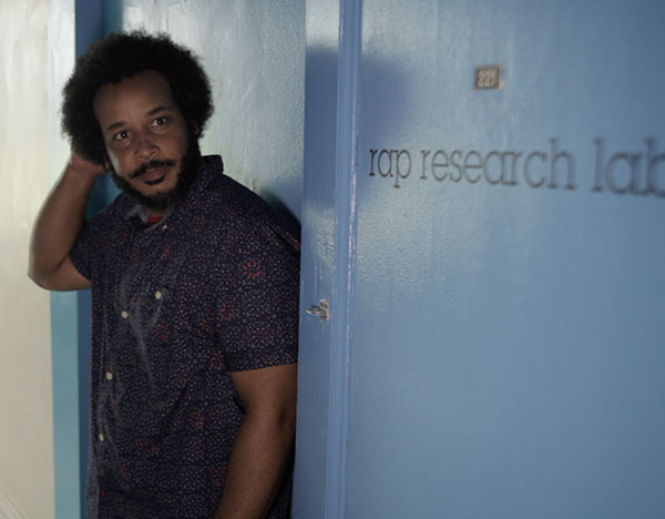 A mid-toned Black man with a mid-length afro, stands in front of a light blue door labeled "rap research lab".