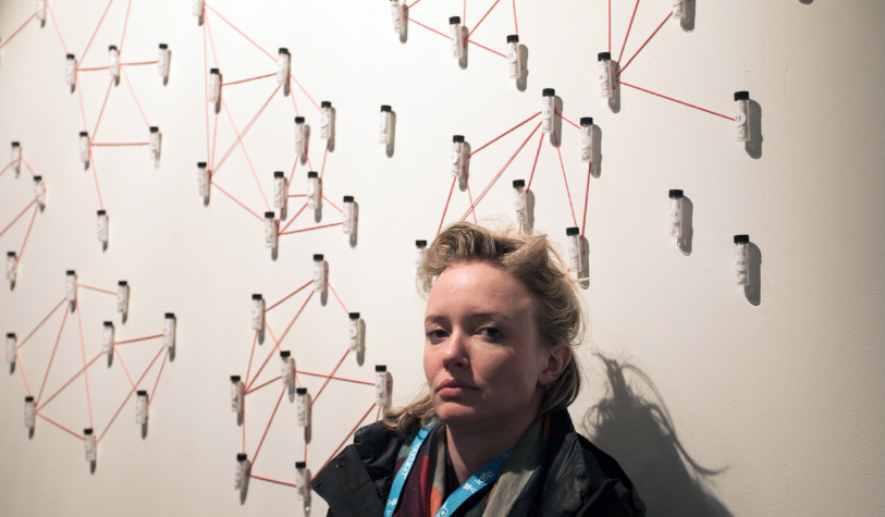 Artist Tega Brain, a White Woman with blond hair, with hair tied back, stands in front of a network visualization. It looks like network visualization is used made with a red thread attached to a white wall.