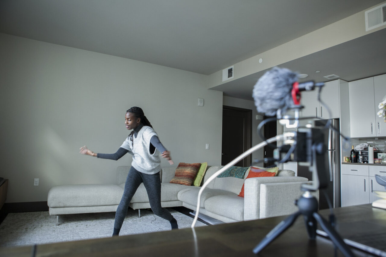Pictured is Valencia James, a Black dark-skinned Dancer, wearing a light gray no-sleeve cable-knitted jumper over a darker gray full body, long-sleeved loose leotard. She stands in with arms apart in a dance pose, being recorded by a handmade volumetric motion capture device (her project). She dances in an empty space of her living room, around a light grey couch.