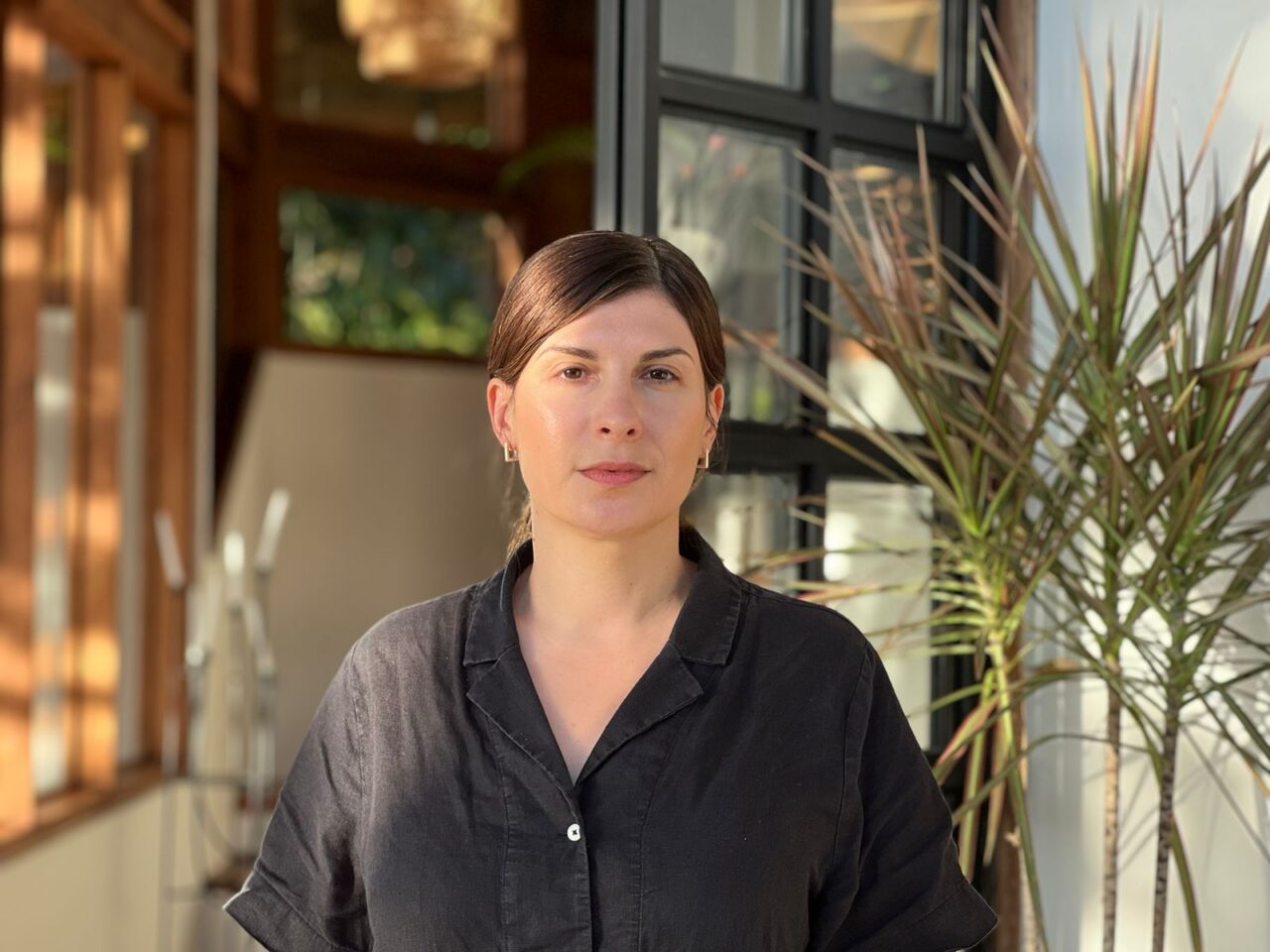 Portrait of Julia Kaganskiy, with medium-length, honeyed brown hair tied back, standing in a light-filled studio space. This picture was taken at Studio Alto, a residency in Costa Rica, nestled on the hilltops of Playa Grande, in a sun-filled room amidst a lush green mountainscape reflected throughout the windows of this space.